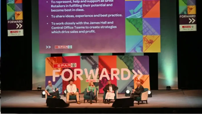 Five panelists sit on stage in armchairs during a SPAR Forward event, with a colorful geometric backdrop and a large screen displaying bullet points above them.