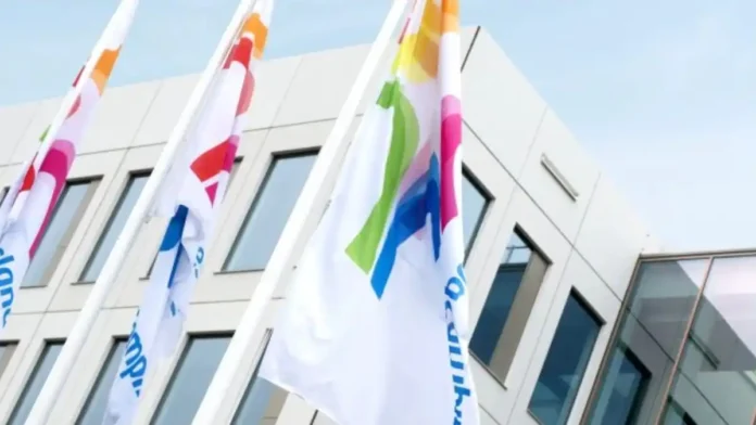White flags with colorful abstract logos flutter in front of a modern office building with large windows and clean geometric lines.